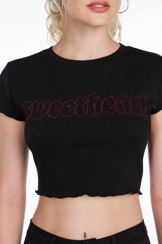 An individual is dressed in the "Sweetheart Lettuce Trim Baby Tee" by Sunshine Rollers, featuring "sweetheart" in a red, decorative font on a black fabric. The top has ruffled edges on the sleeves and hem, set against a plain white background.