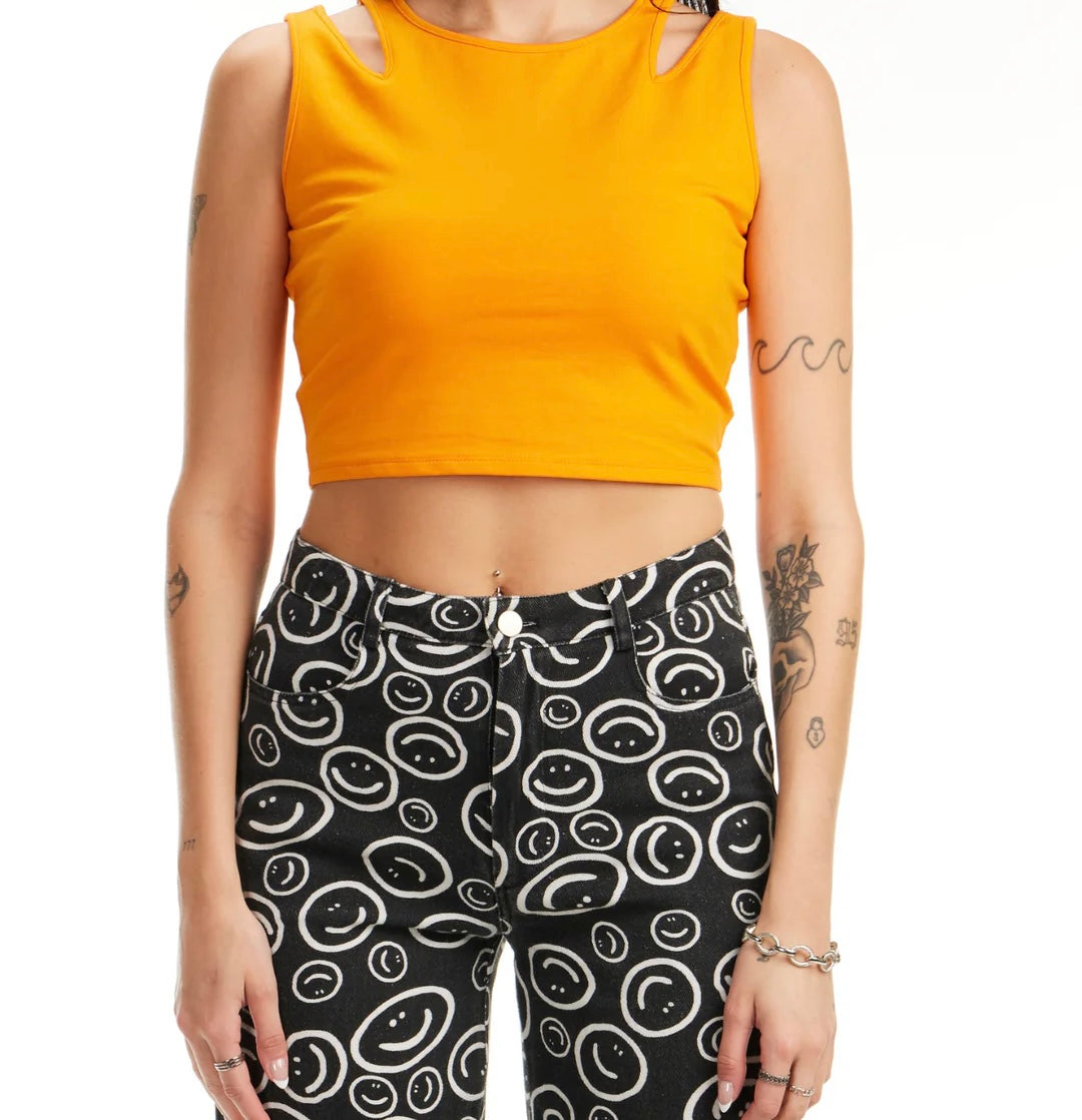 The individual is sporting the Pretty Damage - Orange Tank Top by Sunshine Rollers, paired with black pants featuring a playful white smiley face print. Their arms are adorned with tattoos and stylish bracelets, while their face remains unseen, contributing an air of mystery to their edgy ensemble.