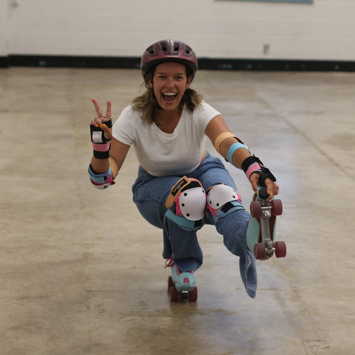 A person wearing a helmet and protective gear skates indoors on roller skates, smiling and making a peace sign while balancing on one foot. Enjoying their Sunshine Rollers from the Open Skate Package (16+) - Nov/Dec 2024 in the empty room with a concrete floor, they're clearly thrilled with their choice from the local skate rental shop.