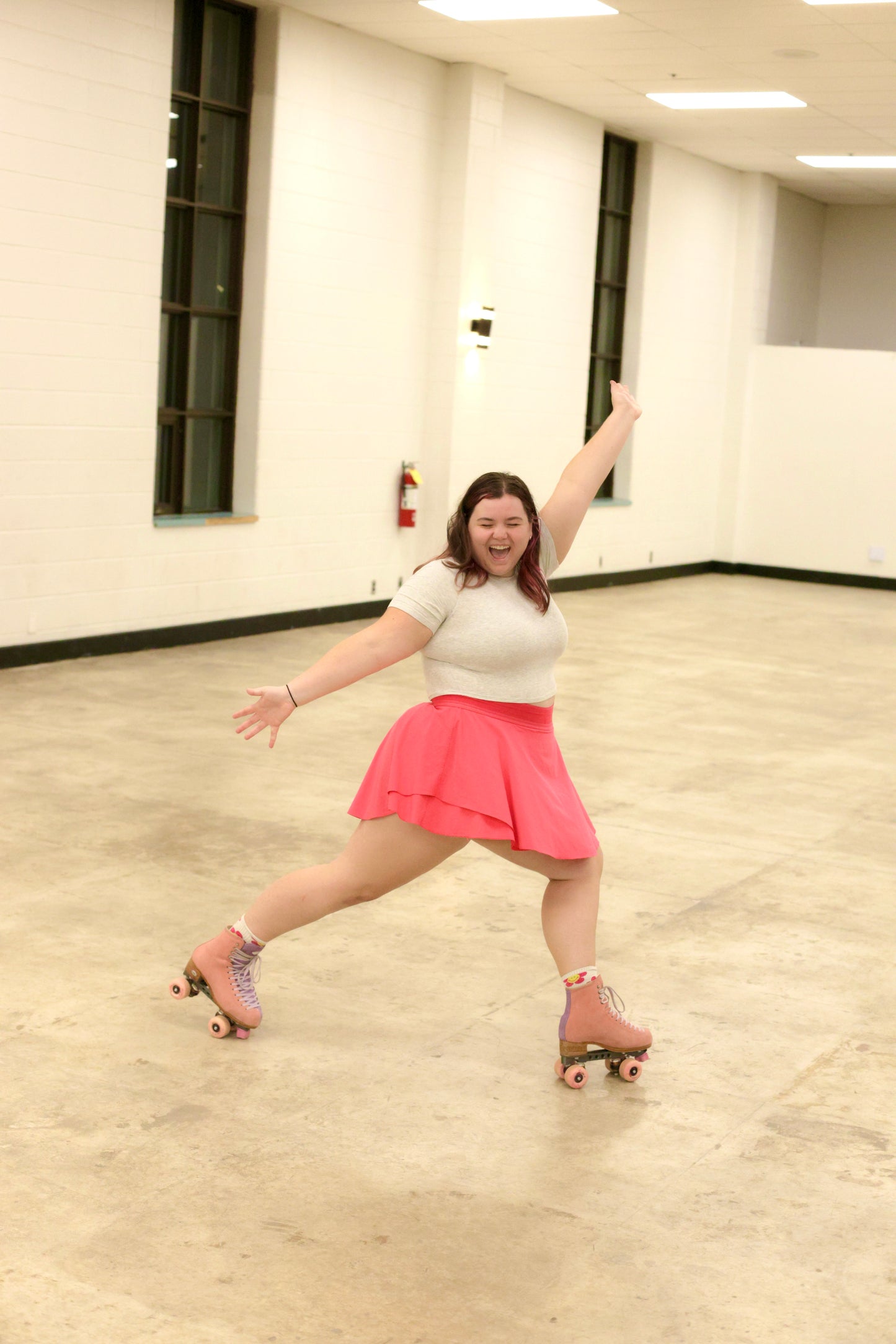 A woman joyfully roller-skates indoors, capturing the excitement of a Roller Dance Workshop with Des by Sunshine Rollers. She's wearing a bright pink skirt and light-colored top, with one arm extended upwards and a broad smile that showcases her energetic and fun spirit. The room is illuminated by large windows and has a polished floor.
