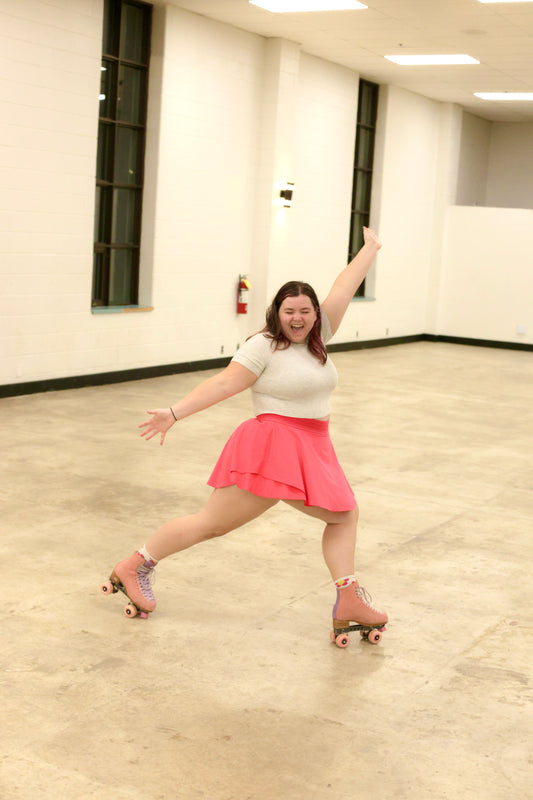 A woman joyfully roller-skates indoors, capturing the excitement of a Roller Dance Workshop with Des by Sunshine Rollers. She's wearing a bright pink skirt and light-colored top, with one arm extended upwards and a broad smile that showcases her energetic and fun spirit. The room is illuminated by large windows and has a polished floor.