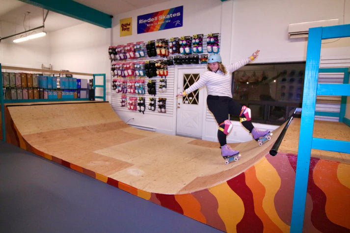 A person wearing roller skates and protective gear participates in a Sunshine Rollers MINI RAMP - 1 Hour Private Lesson on an indoor mini ramp. The room features colorful socks displayed on the wall and a rack of skateboards, while the ramp is designed with vibrant orange and red wave patterns along its sides.