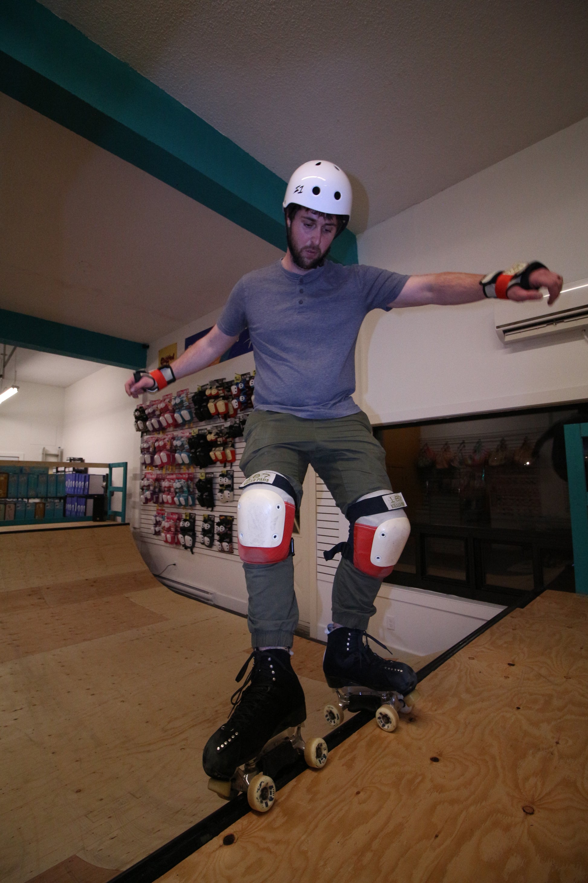 A person clad in a helmet and protective gear is engaged in a one-hour private lesson using the MINI RAMP, offered by Sunshine Rollers, while roller skating indoors. In the background, shelves brimming with skating equipment are visible.