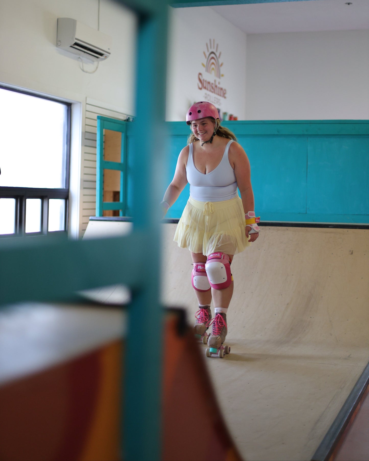 A woman wearing a pink helmet, white tank top, yellow tutu skirt, and pink roller skate pads rides a roller skate up an indoor wooden ramp. The background shows a teal and white wall with a 'Sunshine' logo.