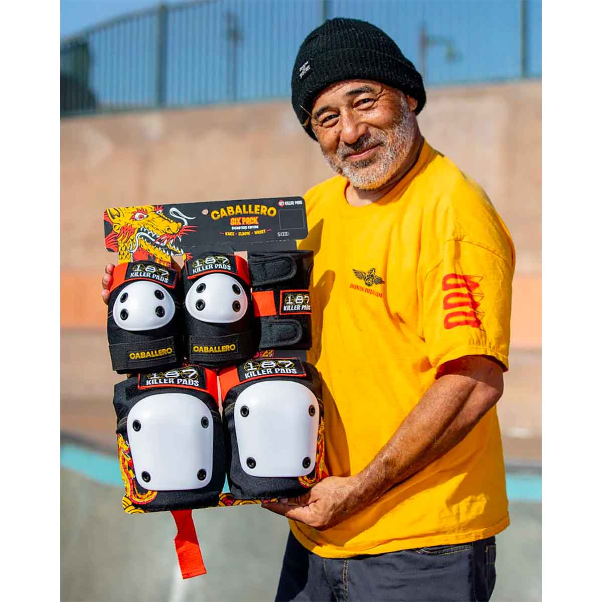 A smiling man wearing a yellow shirt and black beanie holds packages of the 187 Killer Pads - Adult 6 Piece Combo Pack (Steve Caballero) by Sunshine Rollers. He's standing in Skate Park with a blurred ramp in the background, evoking a classic Steve Caballero scene.